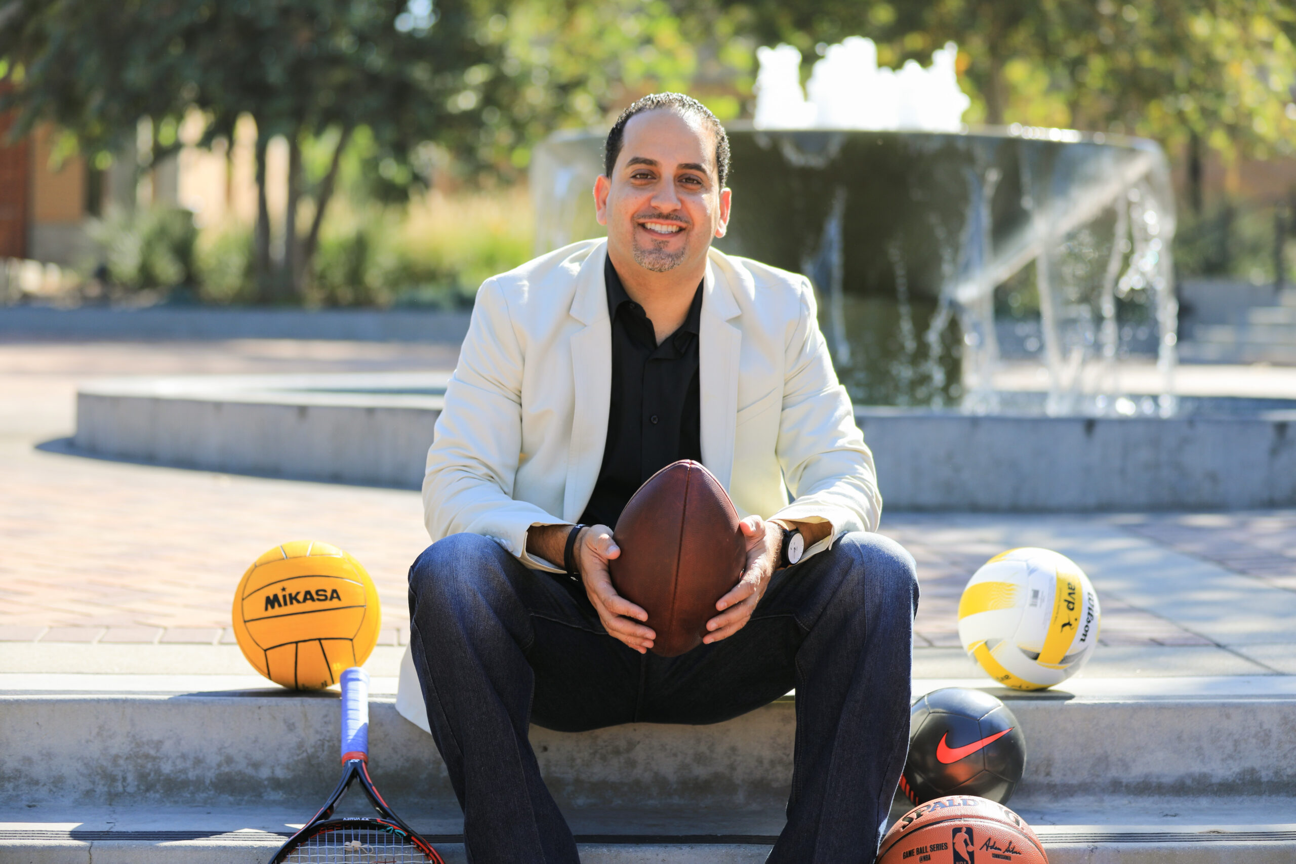 Drew Marcos in front of a fountain gripping a football and surrounded by various sports equipment.
